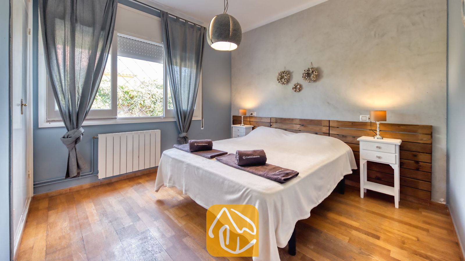 Vakantievilla's Costa Brava Spanje - Villa Yara - Slaapkamer met een tweepersoonsbed, een houten hoofdbord, twee witte nachtkastjes met kleine lampjes, en grijze gordijnen die het raam omlijsten.