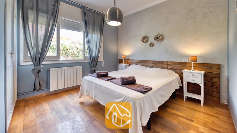 Vakantievilla's Costa Brava Spanje - Villa Yara - Slaapkamer met een tweepersoonsbed, een houten hoofdbord, twee witte nachtkastjes met kleine lampjes, en grijze gordijnen die het raam omlijsten.
