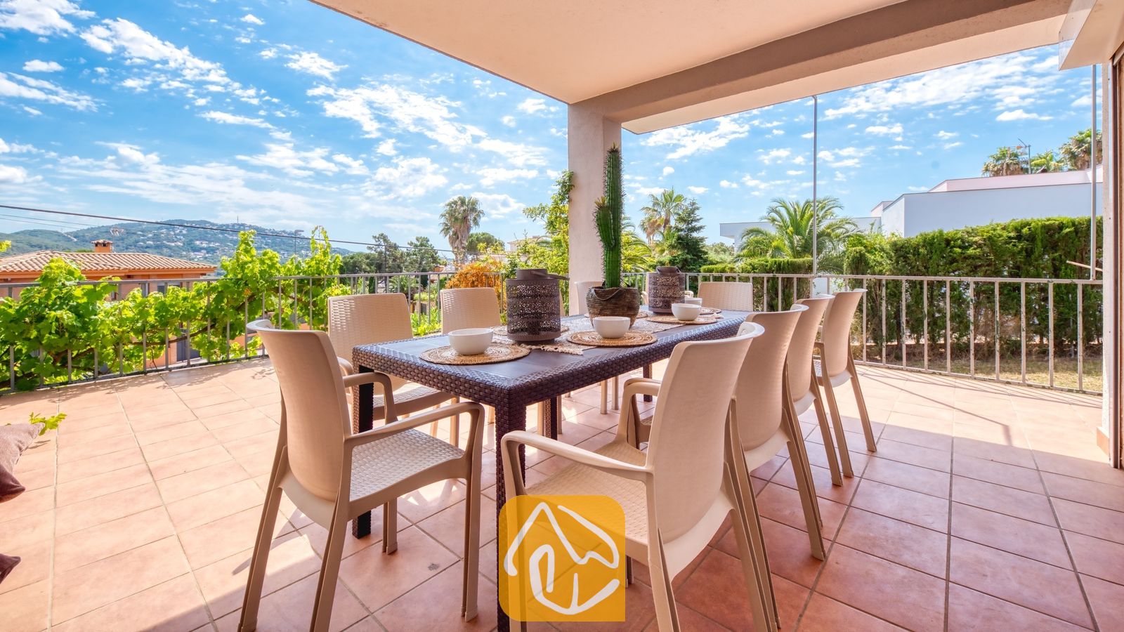 Vakantievilla's Costa Brava Spanje - Villa Yara - Overdekt terras bij Villa Yara, voorzien van een eettafel in de buitenlucht omringd door kunststof stoelen, tafelsetting met kommen en in de verte heuvels onder een blauwe hemel.