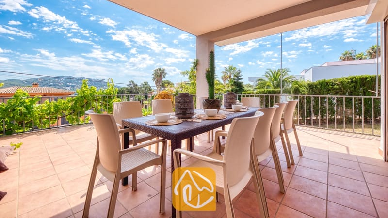Vakantievilla's Costa Brava Spanje - Villa Yara - Overdekt terras bij Villa Yara, voorzien van een eettafel in de buitenlucht omringd door kunststof stoelen, tafelsetting met kommen en in de verte heuvels onder een blauwe hemel.