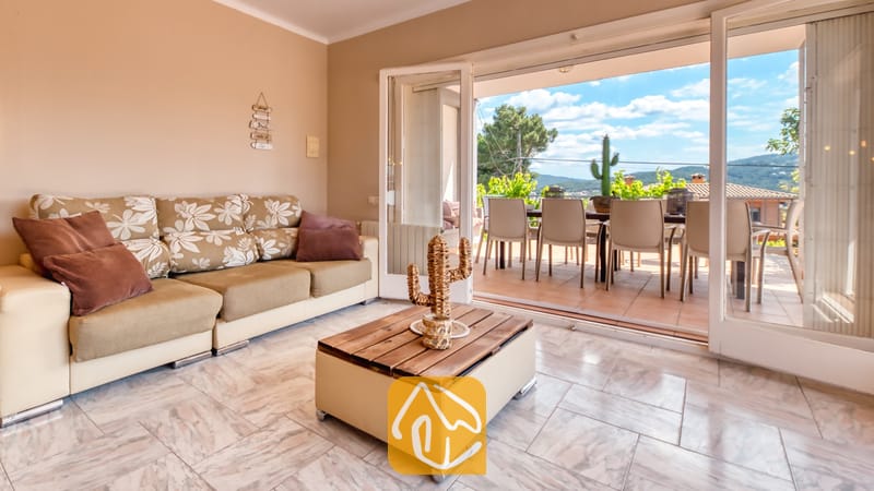 Vakantievilla's Costa Brava Spanje - Villa Yara - Woonkamer van Villa Yara met een bank met bloemenprint, een houten salontafel en open glazen deuren die leiden naar een zonnig terras met eethoek en uitzicht op de heuvels.