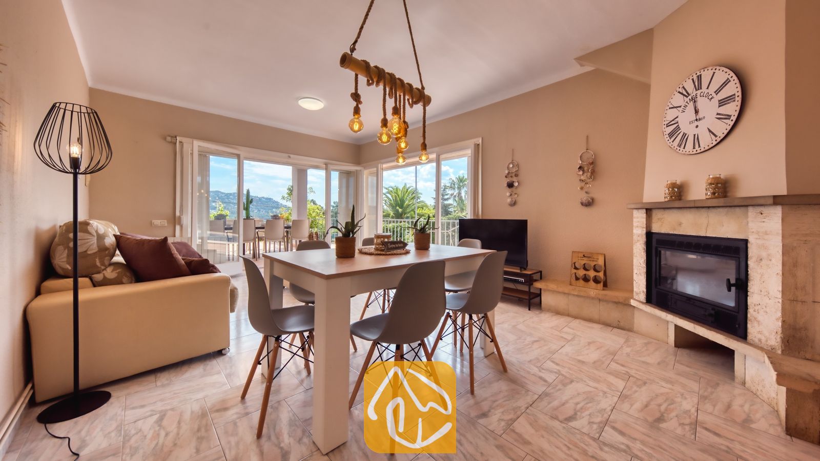 Vakantievilla's Costa Brava Spanje - Villa Yara - Woon- en eethoek in Villa Yara, voorzien van een houten eettafel met grijze stoelen, een stenen open haard en grote glazen deuren die uitkomen op een terras.