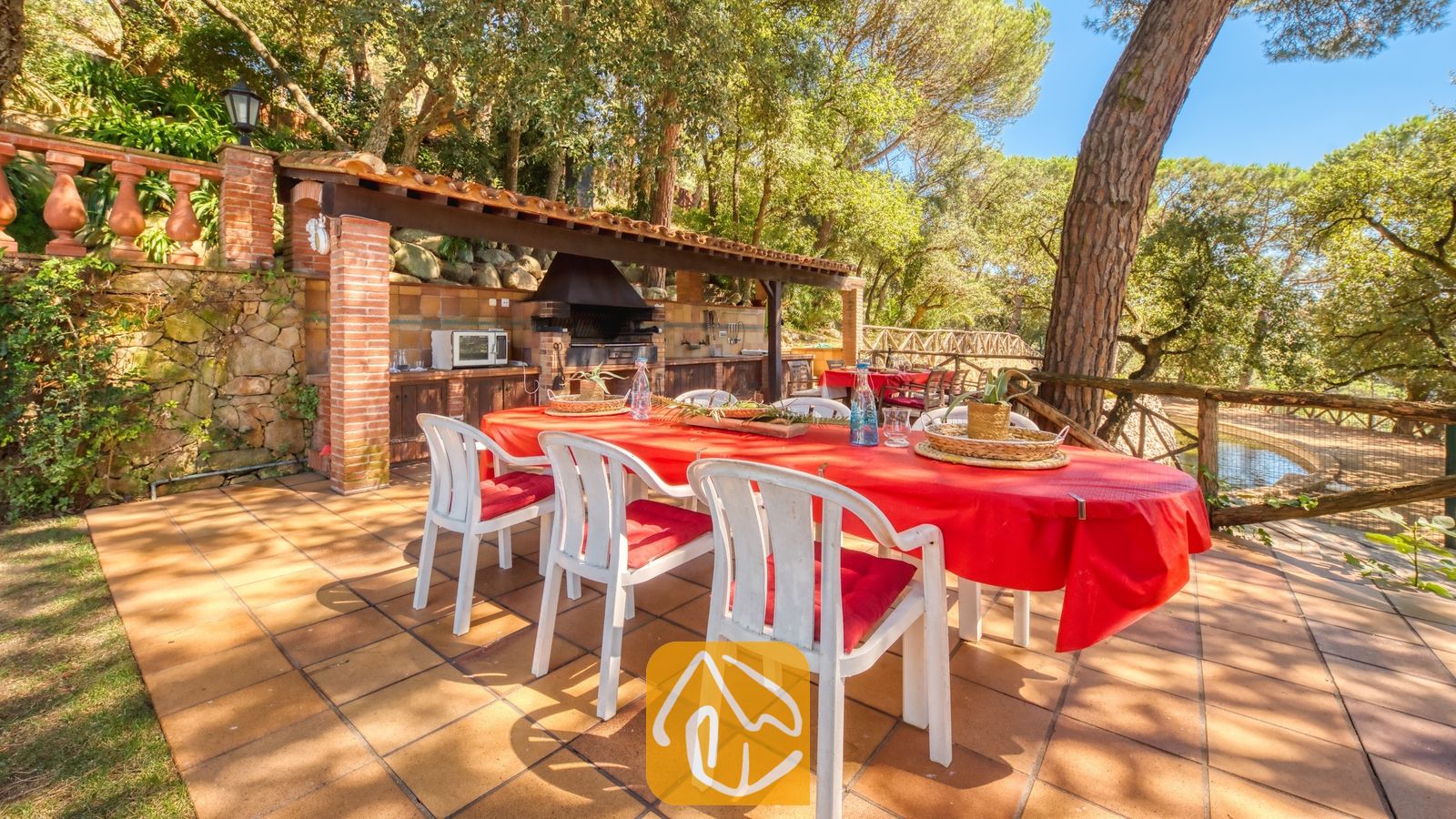 Vakantievilla's Costa Brava Spanje - Villa Paradise - Eethoek in de open lucht bij Villa Paradise, met een grote tafel voorzien van een felrood tafelkleed, witte kunststof stoelen met rode kussens en een ingebouwde stenen barbecue onder een houten overkapping.