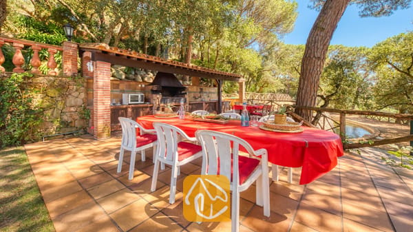 Vakantievilla's Costa Brava Spanje - Villa Paradise - Eethoek in de open lucht bij Villa Paradise, met een grote tafel voorzien van een felrood tafelkleed, witte kunststof stoelen met rode kussens en een ingebouwde stenen barbecue onder een houten overkapping.