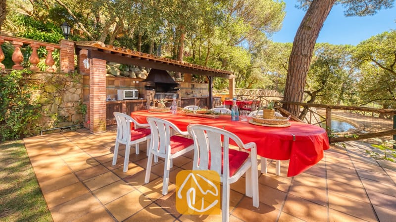 Vakantievilla's Costa Brava Spanje - Villa Paradise - Eethoek in de open lucht bij Villa Paradise, met een grote tafel voorzien van een felrood tafelkleed, witte kunststof stoelen met rode kussens en een ingebouwde stenen barbecue onder een houten overkapping.