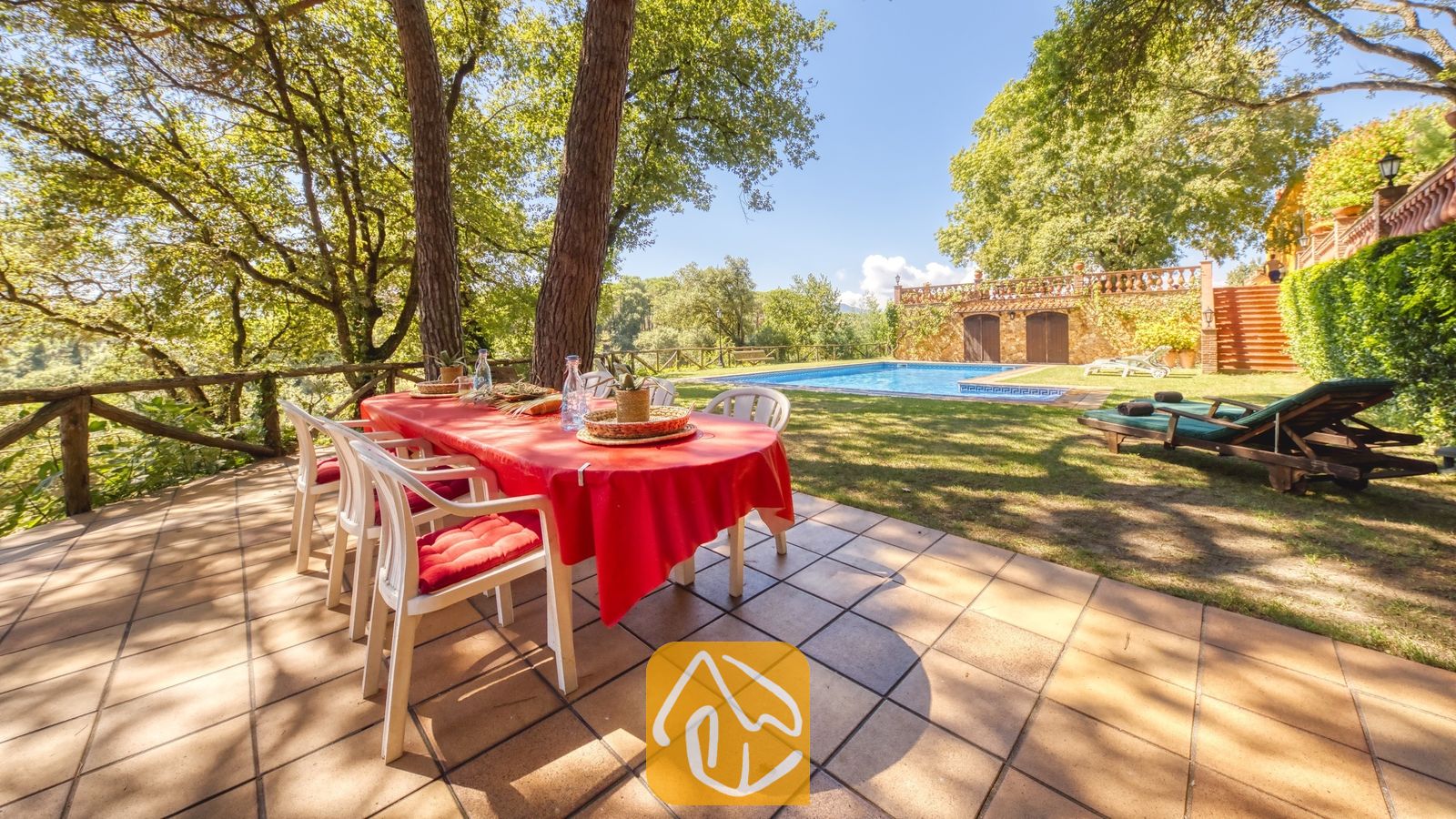 Vakantievilla's Costa Brava Spanje - Villa Paradise - Buiteneettafel met een opvallend rood tafelkleed op een stenen terras bij Villa Paradise, met uitzicht op een groen gazon, een zwembad en bomen.