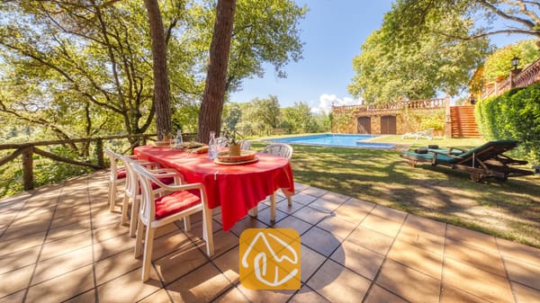 Vakantievilla's Costa Brava Spanje - Villa Paradise - Buiteneettafel met een opvallend rood tafelkleed op een stenen terras bij Villa Paradise, met uitzicht op een groen gazon, een zwembad en bomen.