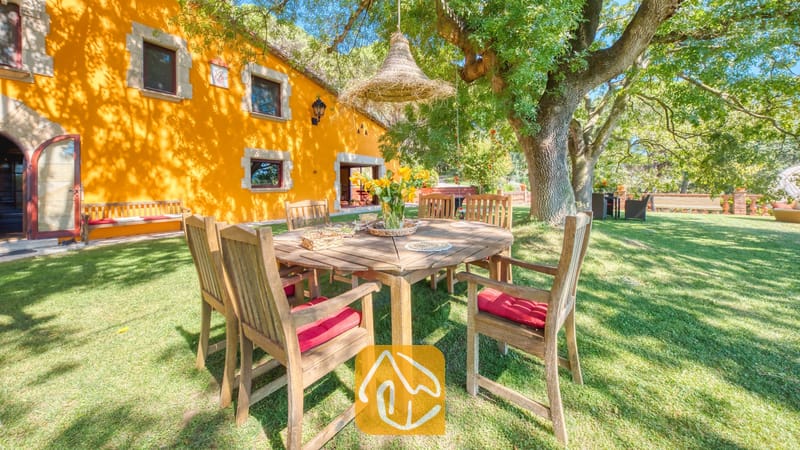 Vakantievilla's Costa Brava Spanje - Villa Paradise - Houten eethoek met rode kussens op een groen gazon naast Villa Paradise, met heldergele buitenmuren en grote schaduwrijke bomen.