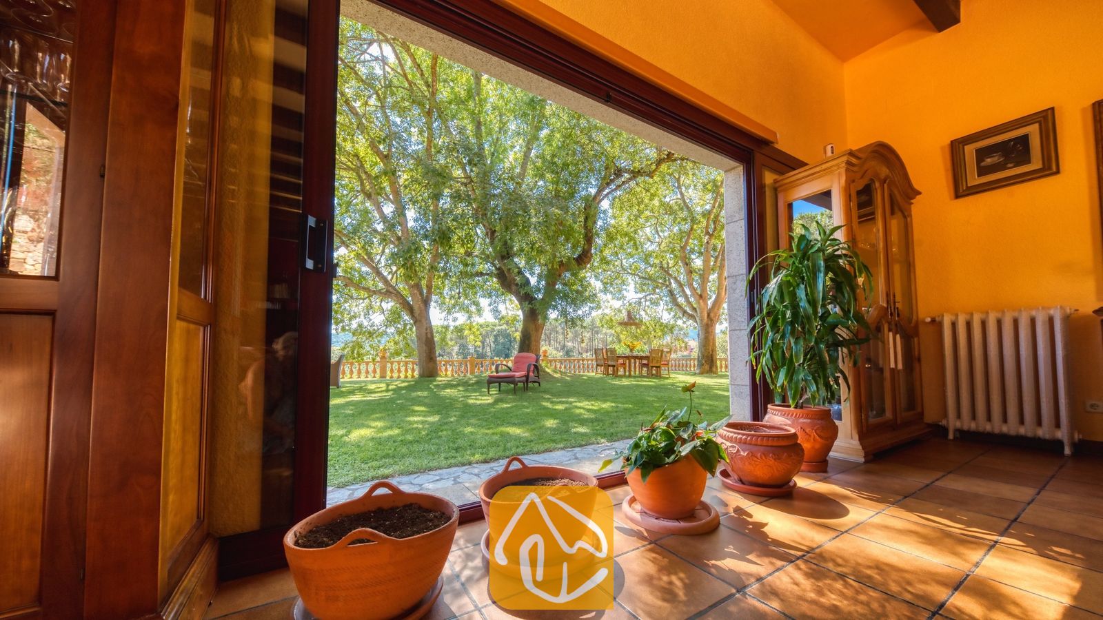 Vakantievilla's Costa Brava Spanje - Villa Paradise - Open de glazen schuifdeur van Villa Paradise en u kijkt uit op een groen gazon met grote bomen, een betegeld terras en sfeervolle potplanten.