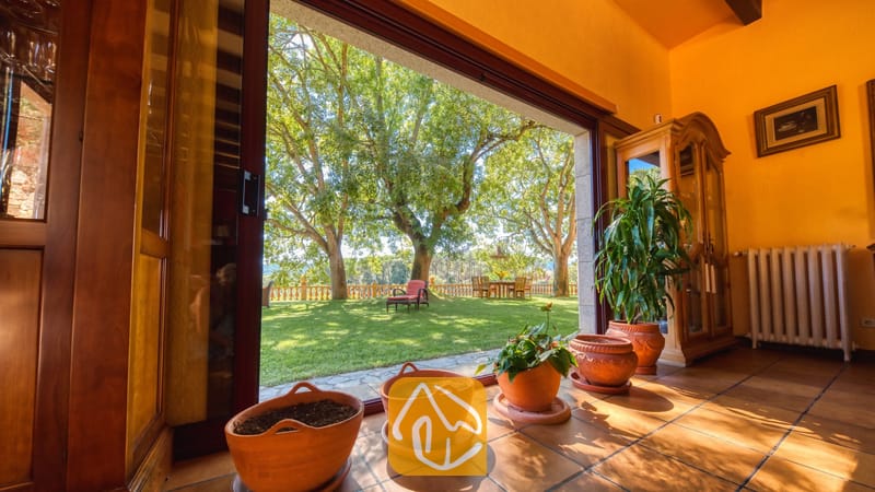 Vakantievilla's Costa Brava Spanje - Villa Paradise - Open de glazen schuifdeur van Villa Paradise en u kijkt uit op een groen gazon met grote bomen, een betegeld terras en sfeervolle potplanten.