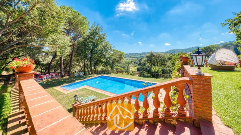Vakantievilla's Costa Brava Spanje - Villa Paradise - Privézwembad bij Villa Paradise, omringd door een groen gazon, hoge dennenbomen en een stenen trap met terracotta balustrades die vanaf het terras naar beneden leidt onder een blauwe hemel.