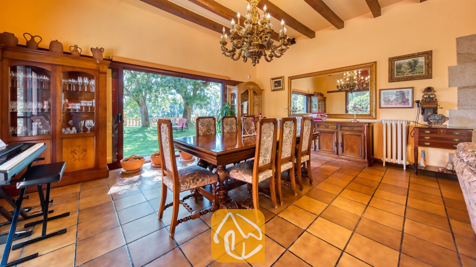 Vakantievilla's Costa Brava Spanje - Villa Paradise - Traditionele eetkamer in Villa Paradise met een grote houten tafel, stoelen met motief, een sierlijke kroonluchter, zichtbare plafondbalken en schuifpui die uitkomt op een tuin.