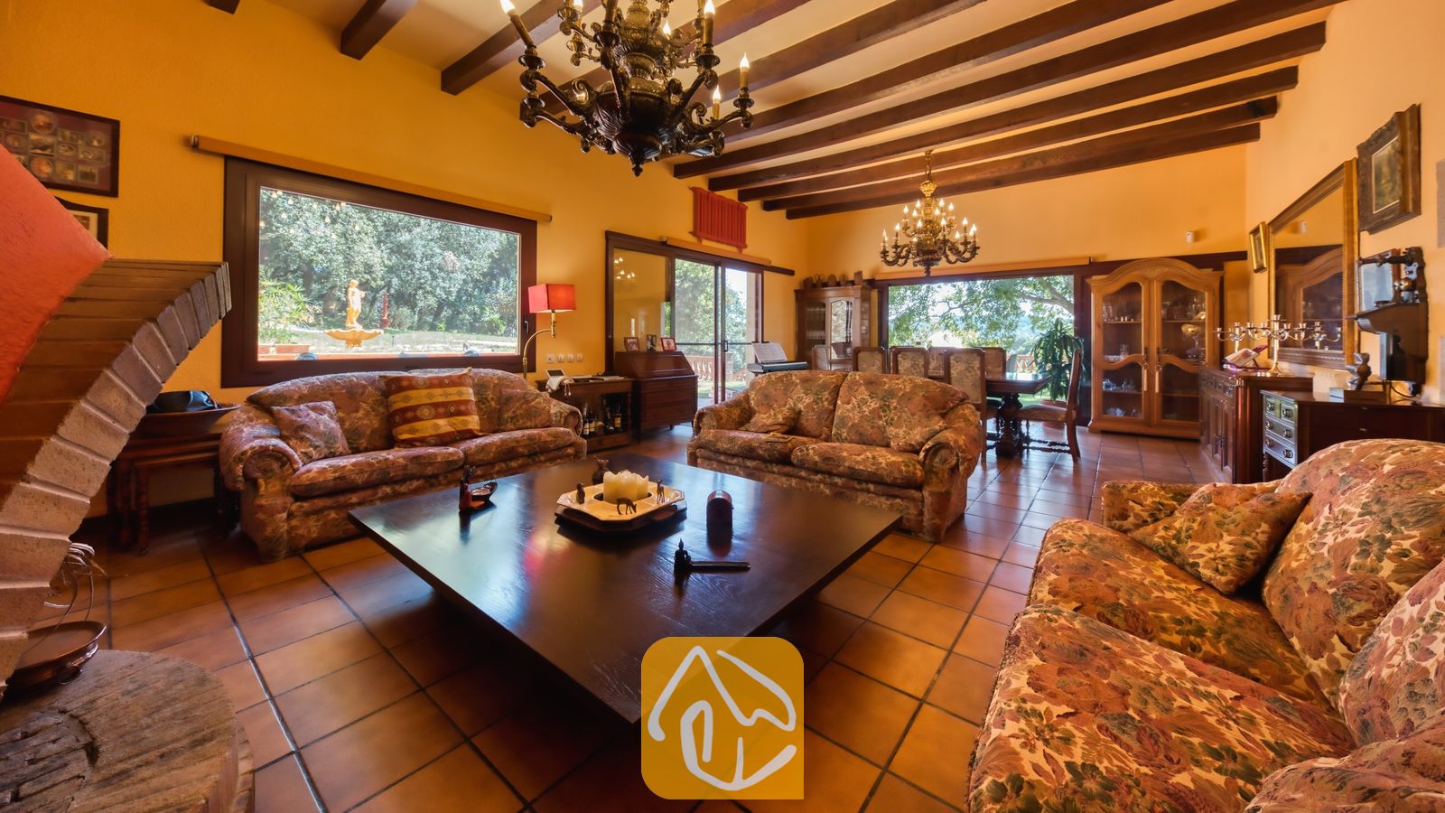Vakantievilla's Costa Brava Spanje - Villa Paradise - Ruime woonkamer in Villa Paradise met authentieke houten plafondbalken, sfeervolle banken met patroon, een grote salontafel van donker hout, tegelvloeren en een stenen open haard.