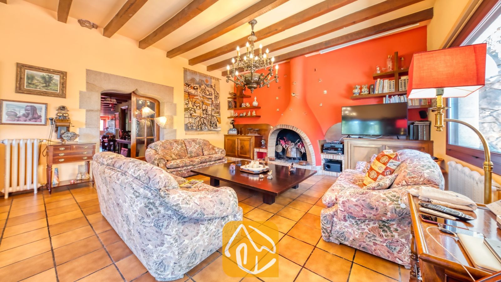 Vakantievilla's Costa Brava Spanje - Villa Paradise - Woonkamer van Villa Paradise met een oranje accentmuur, een stenen open haard, zichtbare houten plafondbalken en drie banken met een geprint stoffen patroon.