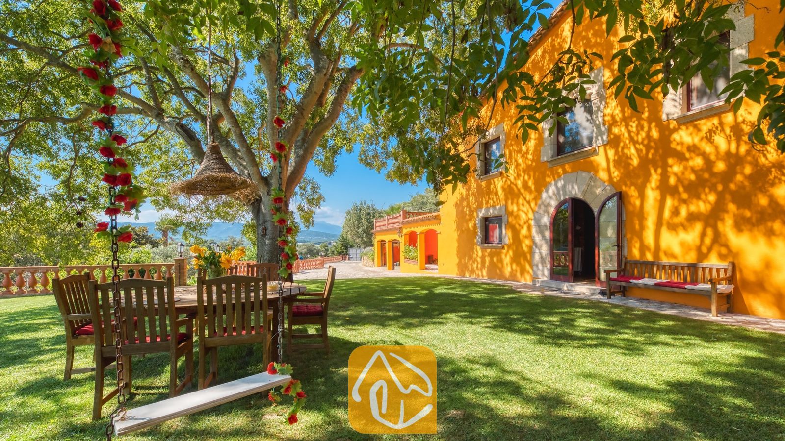 Vakantievilla's Costa Brava Spanje - Villa Paradise - Zonnige tuin bij Villa Paradise met een houten schommel versierd met rode bloemen, een eettafel buiten onder een grote boom en de heldergele buitenmuur van de villa.