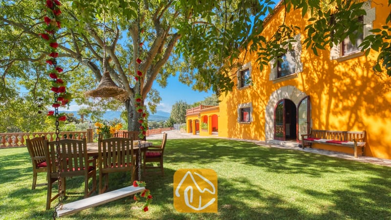 Vakantievilla's Costa Brava Spanje - Villa Paradise - Zonnige tuin bij Villa Paradise met een houten schommel versierd met rode bloemen, een eettafel buiten onder een grote boom en de heldergele buitenmuur van de villa.