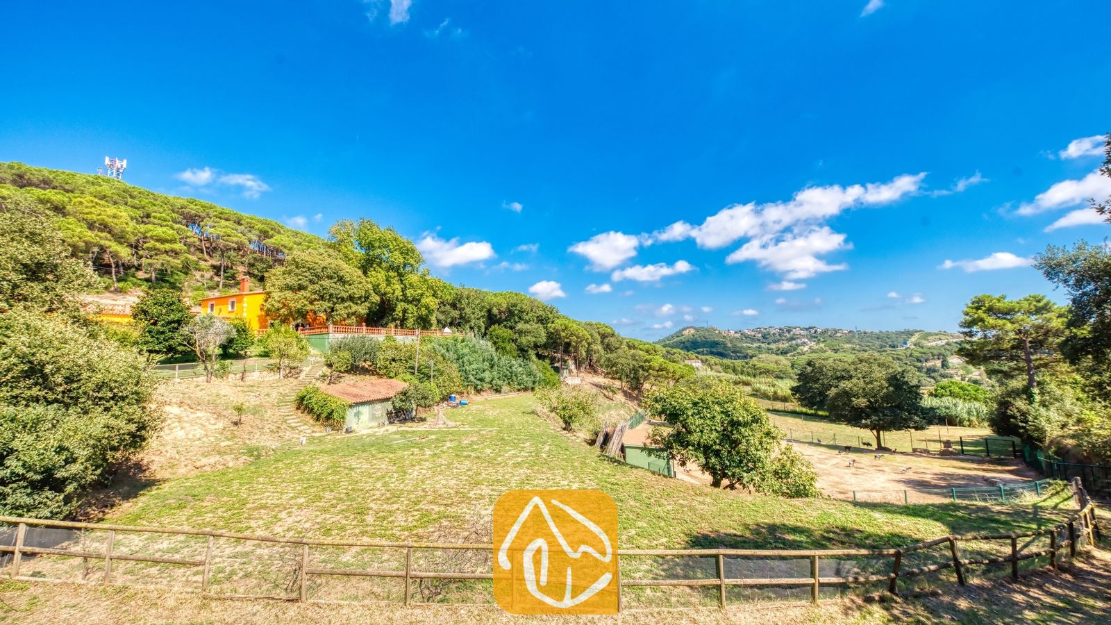 Vakantievilla's Costa Brava Spanje - Villa Paradise - Buitenaanzicht van Villa Paradise, met een oranje gebouw verscholen tussen groene heuvels, omheinde grasvelden en een blauwe lucht met her en der wat wolken.