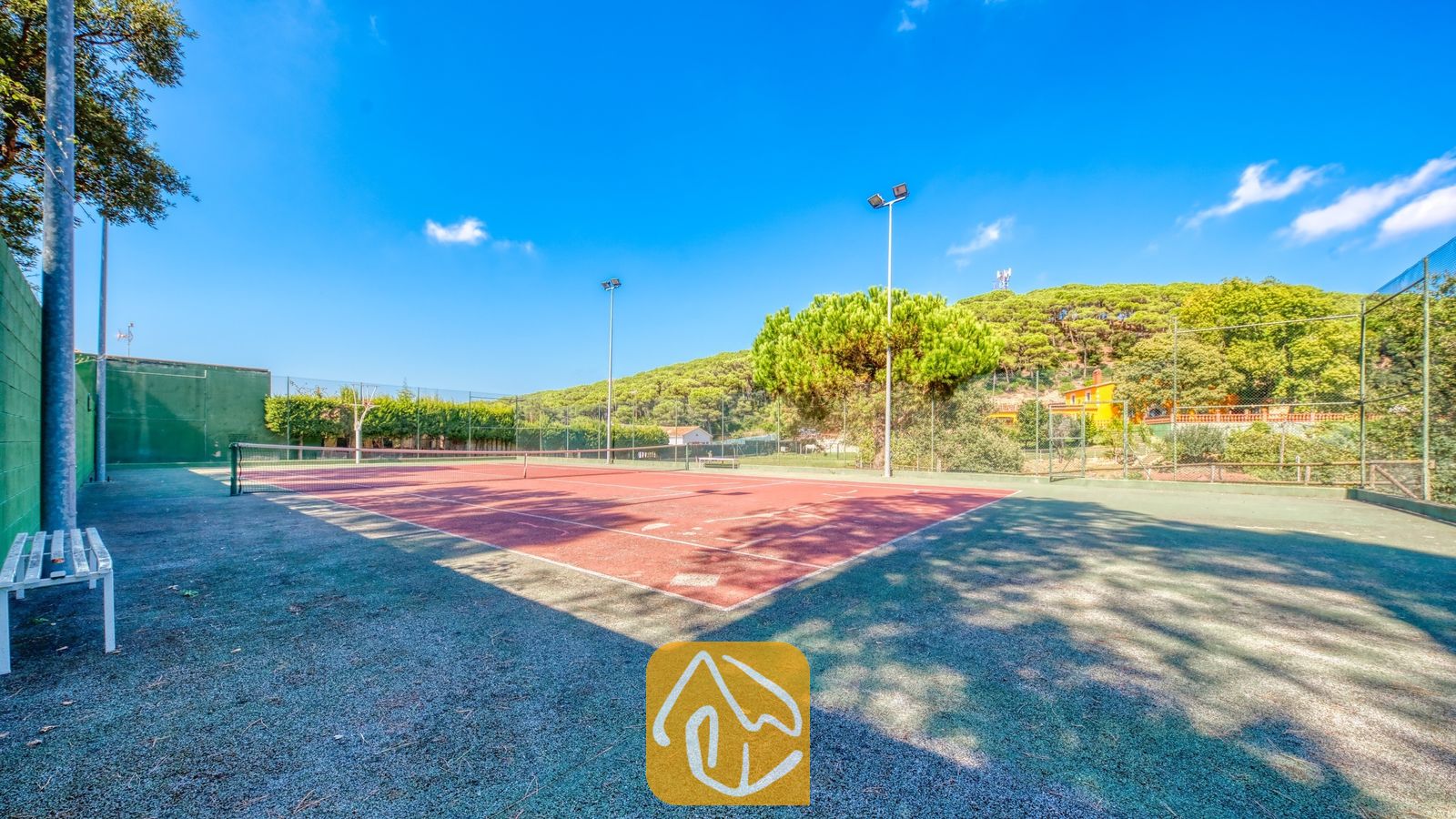 Vakantievilla's Costa Brava Spanje - Villa Paradise - Tennisbaan bij Villa Paradise met een rode ondergrond, witte belijningslijnen, net en een omringend groen schakelkettinghek onder een blauwe lucht.