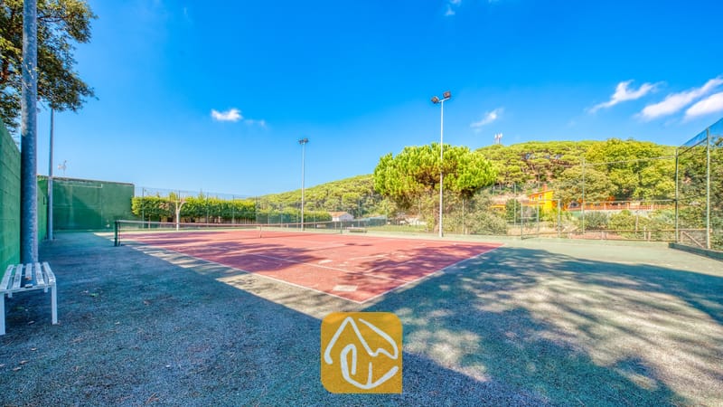Vakantievilla's Costa Brava Spanje - Villa Paradise - Tennisbaan bij Villa Paradise met een rode ondergrond, witte belijningslijnen, net en een omringend groen schakelkettinghek onder een blauwe lucht.