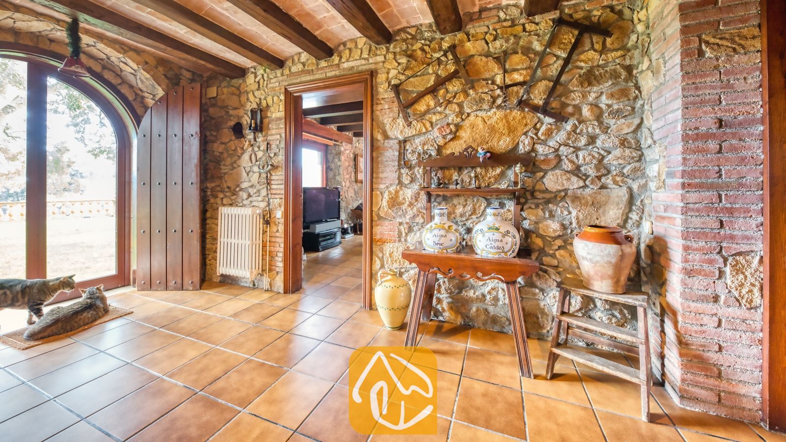 Vakantievilla's Costa Brava Spanje - Villa Paradise - Rustieke muren van natuursteen met zichtbare houten balken aan het plafond, terracotta plavuizen, twee katten bij een boogvormige glazen deur en traditioneel aardewerk op houten standaards.