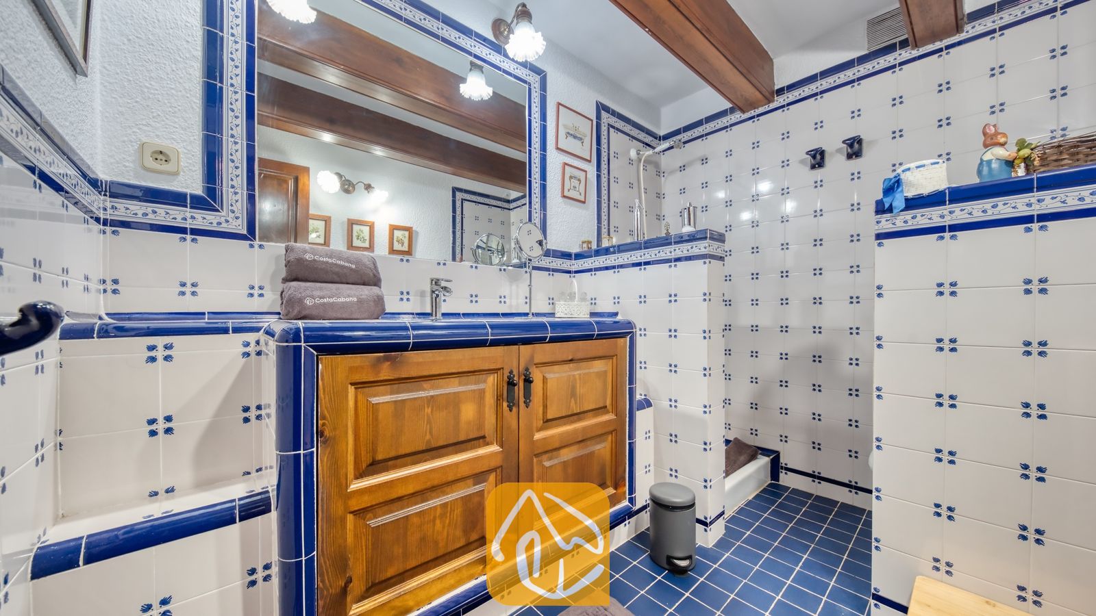 Vakantievilla's Costa Brava Spanje - Villa Paradise - Badkamer met wit-en-blauwe keramische tegels met motief, een houten badkamermeubel met opgevouwen handdoeken en een inloopdouche.