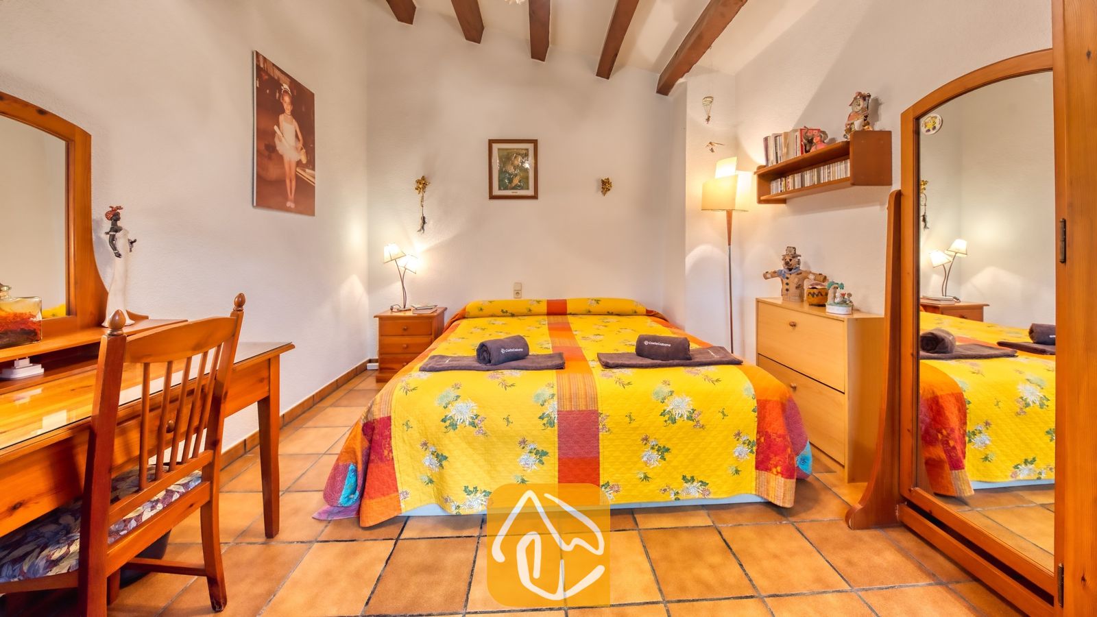 Vakantievilla's Costa Brava Spanje - Villa Paradise - Slaapkamer met een tweepersoonsbed met een geel bloemendekbed, houten meubels waaronder een kaptafel met stoel en authentieke houten balken aan het plafond.