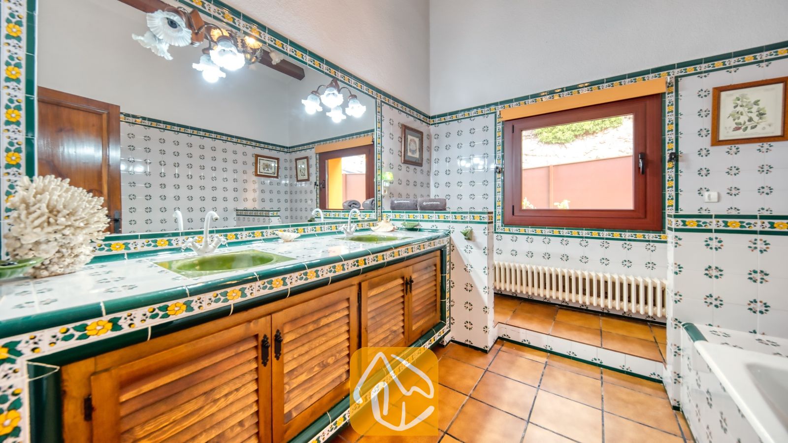 Vakantievilla's Costa Brava Spanje - Villa Paradise - Badmeubel met houten louvredeurtjes en groene wastafels in een tegelblad met bloemmotieven, een grote spiegel en terracotta vloertegels.