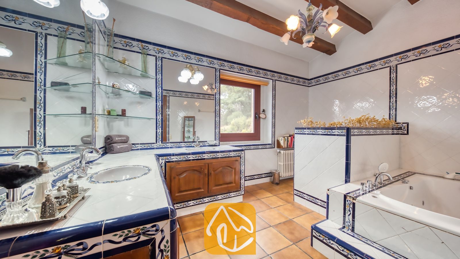 Vakantievilla's Costa Brava Spanje - Villa Paradise - Badkamer met blauw-witte patronen op tegelwerk, een ingebouwd bad, houten kasten en glazen planken tegen witte muren.