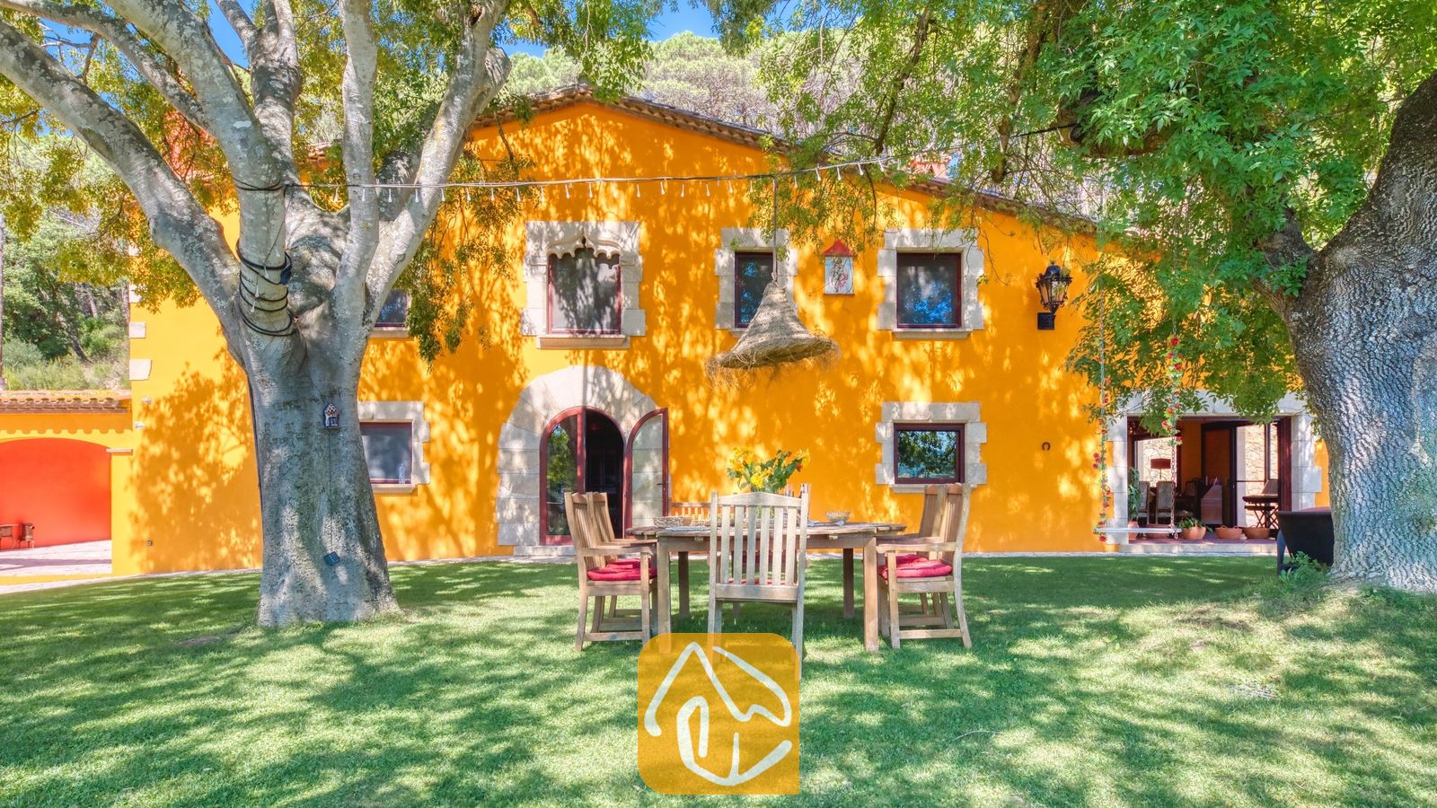 Vakantievilla's Costa Brava Spanje - Villa Paradise - De heldergele buitengevel van Villa Paradise in Spanje, met een houten eettafel buiten op een groen gazon onder grote, volwassen bomen.