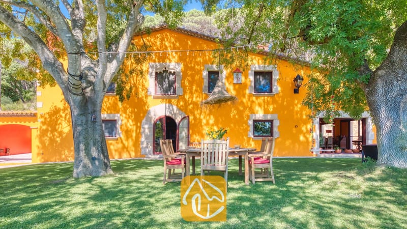 Vakantievilla's Costa Brava Spanje - Villa Paradise - De heldergele buitengevel van Villa Paradise in Spanje, met een houten eettafel buiten op een groen gazon onder grote, volwassen bomen.