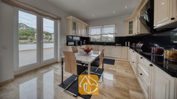 Vakantievilla's Costa Brava Spanje - Villa Madison - Ruime keuken in Villa Madison, voorzien van crèmekleurige kasten, zwarte granieten aanrechtbladen, een glazen eettafel met houten stoelen en dubbele deuren die openen naar een terras.