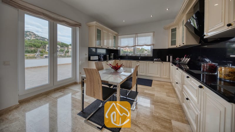 Vakantievilla's Costa Brava Spanje - Villa Madison - Ruime keuken in Villa Madison, voorzien van crèmekleurige kasten, zwarte granieten aanrechtbladen, een glazen eettafel met houten stoelen en dubbele deuren die openen naar een terras.