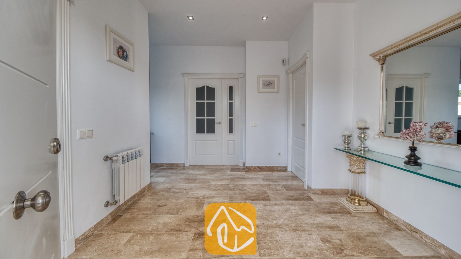 Vakantievilla's Costa Brava Spanje - Villa Madison - Lichte hal met beige stenen plavuizen, witte muren en een glazen sidetable met een decoratieve spiegel.