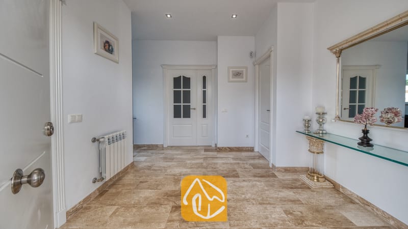 Vakantievilla's Costa Brava Spanje - Villa Madison - Lichte hal met beige stenen plavuizen, witte muren en een glazen sidetable met een decoratieve spiegel.