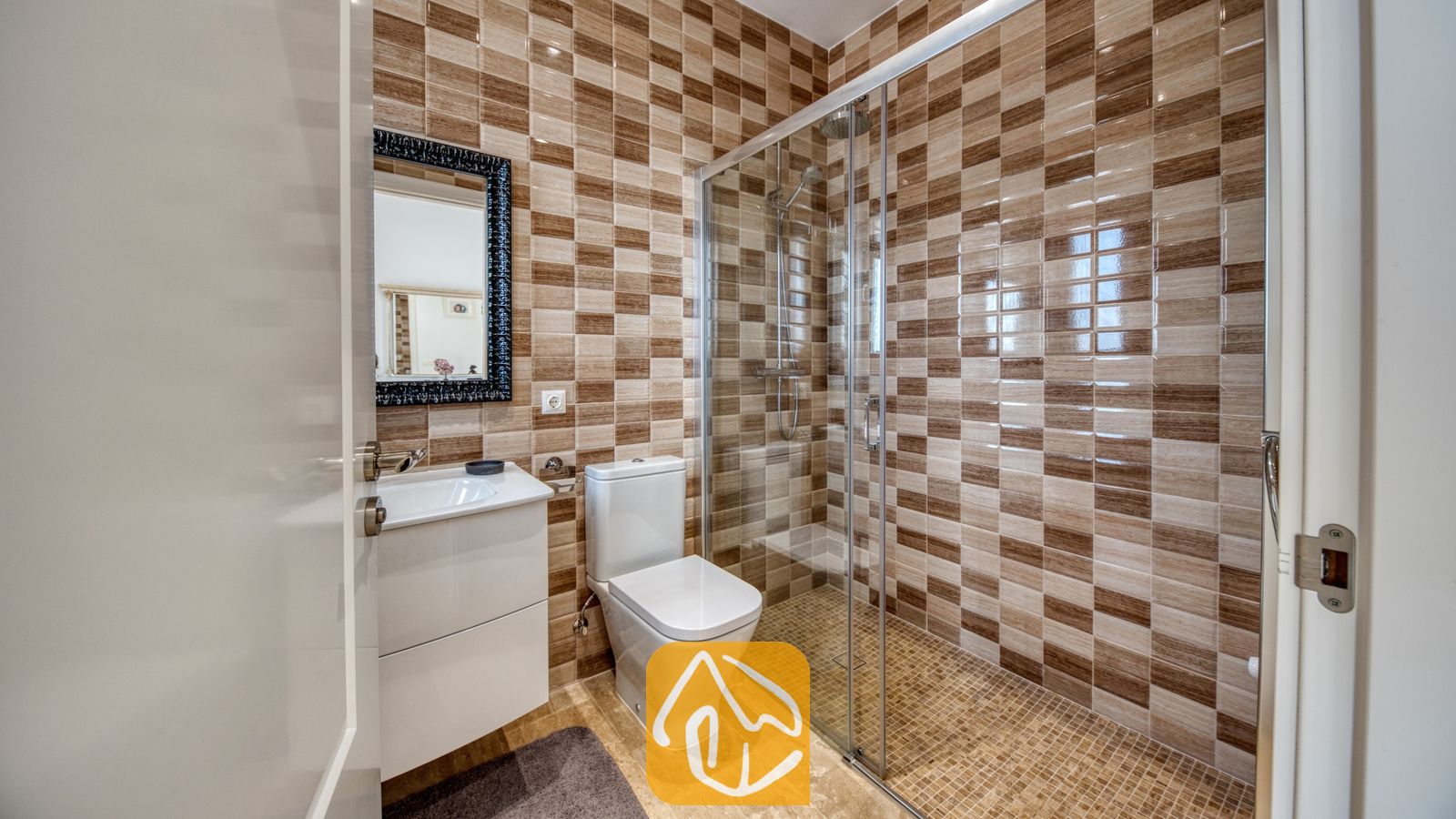 Vakantievilla's Costa Brava Spanje - Villa Madison - Badkamer voorzien van bruine en beige tegels met een dambordmotief, een glazen doucheruimte, een wit toilet en een wastafelmeubel met een zwart ingelijste spiegel.