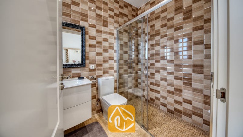 Holiday villas Costa Brava Spain - Villa Madison - Bathroom featuring brown and beige checkerboard wall tiles, a glass-enclosed shower, a white toilet, and a sink vanity with a black-framed mirror.