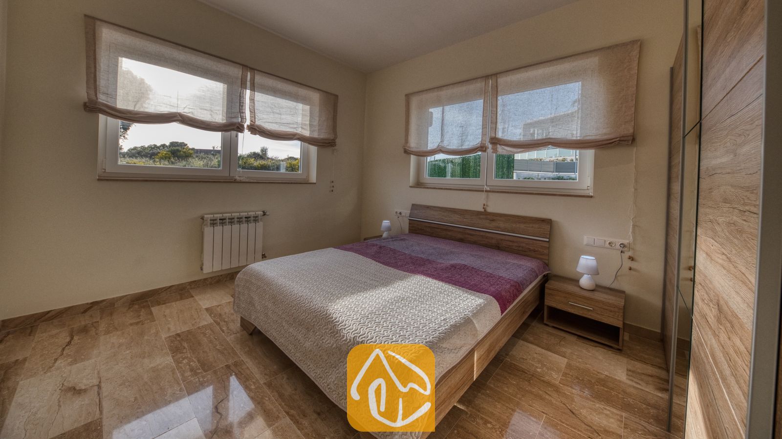 Vakantievilla's Costa Brava Spanje - Villa Madison - Slaapkamer met houten bedframe, twee kleine nachtkastlampjes en grote ramen met stoffen jaloezieën in Villa Madison.