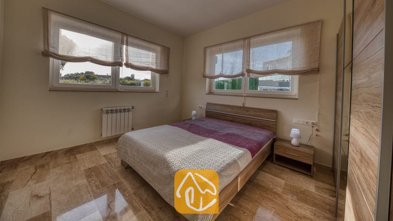 Holiday villas Costa Brava Spain - Villa Madison - Bedroom with wooden bed frame, two small bedside lamps, and large windows with fabric shades at Villa Madison.