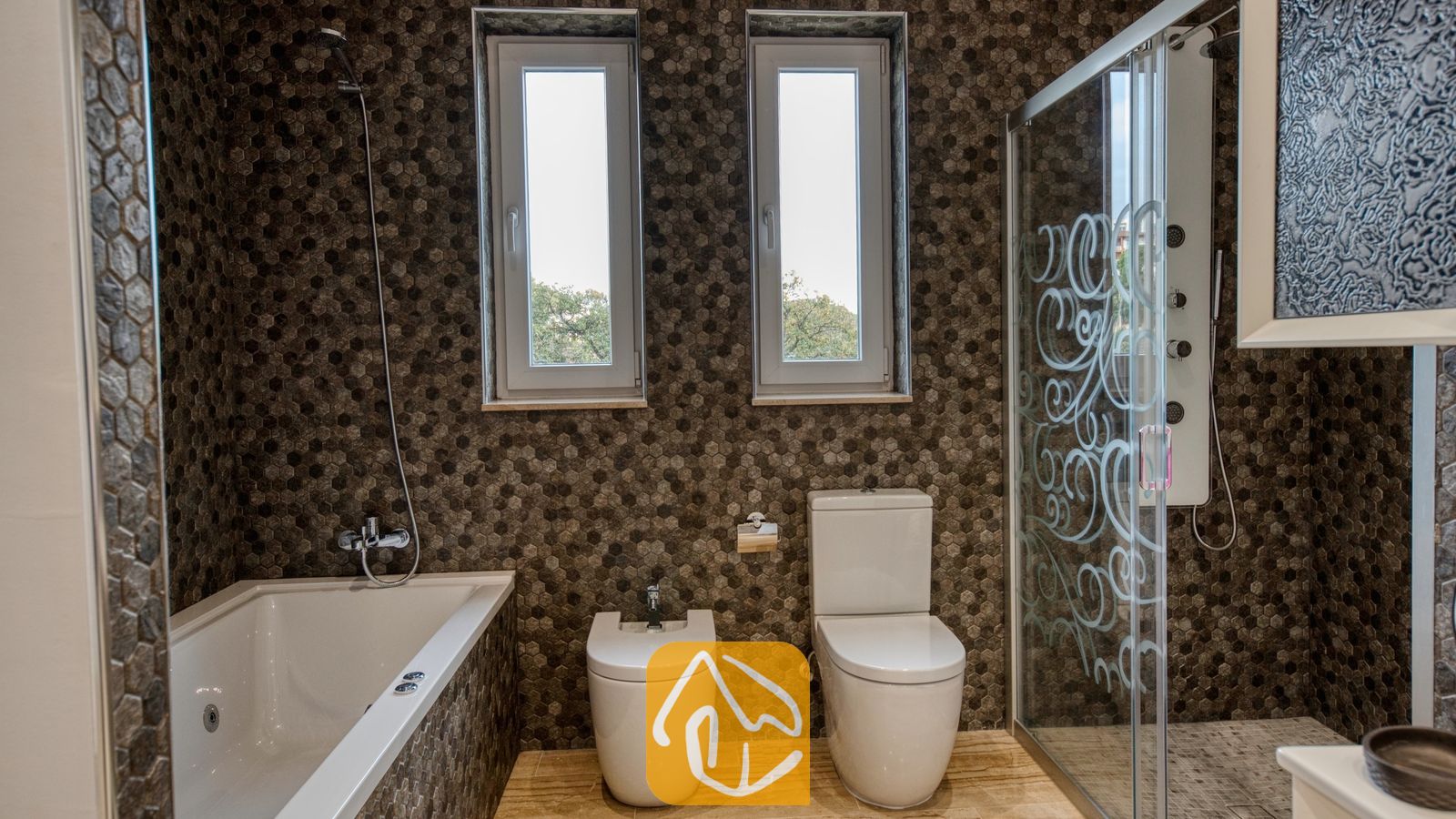 Vakantievilla's Costa Brava Spanje - Villa Madison - Moderne badkamer voorzien van muren met bruine zeshoekige tegels, een wit ligbad, toilet, bidet en een aparte glazen douchecabine.