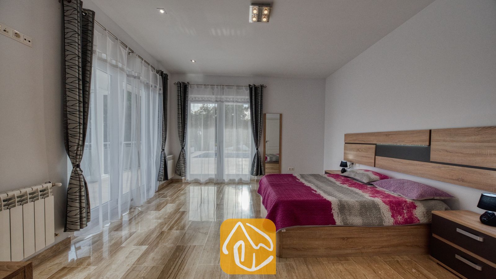 Vakantievilla's Costa Brava Spanje - Villa Madison - Ruime slaapkamer voorzien van een groot houten bed met een roze en grijs geprint sprei, glanzende tegelvloer en glazen schuifdeuren bedekt met doorschijnende witte gordijnen.