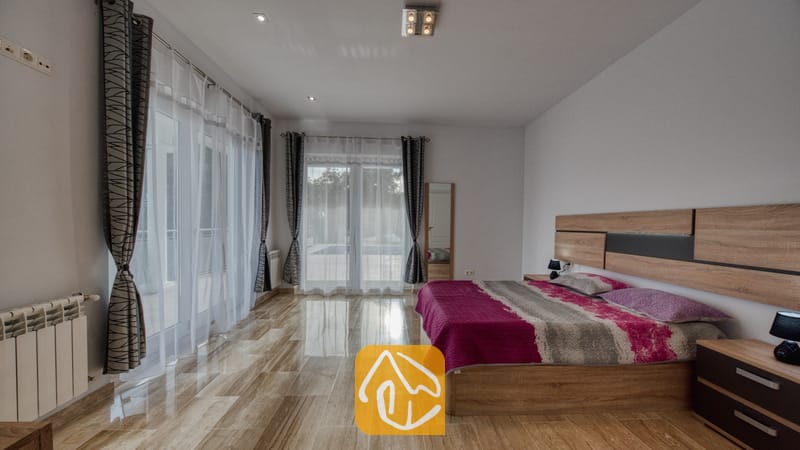 Vakantievilla's Costa Brava Spanje - Villa Madison - Ruime slaapkamer voorzien van een groot houten bed met een roze en grijs geprint sprei, glanzende tegelvloer en glazen schuifdeuren bedekt met doorschijnende witte gordijnen.