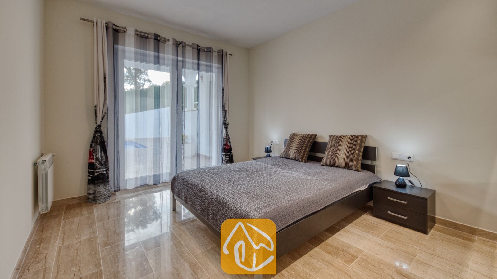 Vakantievilla's Costa Brava Spanje - Villa Madison - Slaapkamer met een tweepersoonsbed, een grijs patroonovertrek, gestreepte kussens en donkere nachtkastjes met lampen, en een glazen deur die toegang biedt tot een terras.