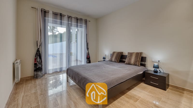 Holiday villas Costa Brava Spain - Villa Madison - Bedroom with double bed, grey patterned cover, striped pillows, and dark nightstands with lamps, featuring a glass door leading to a patio.