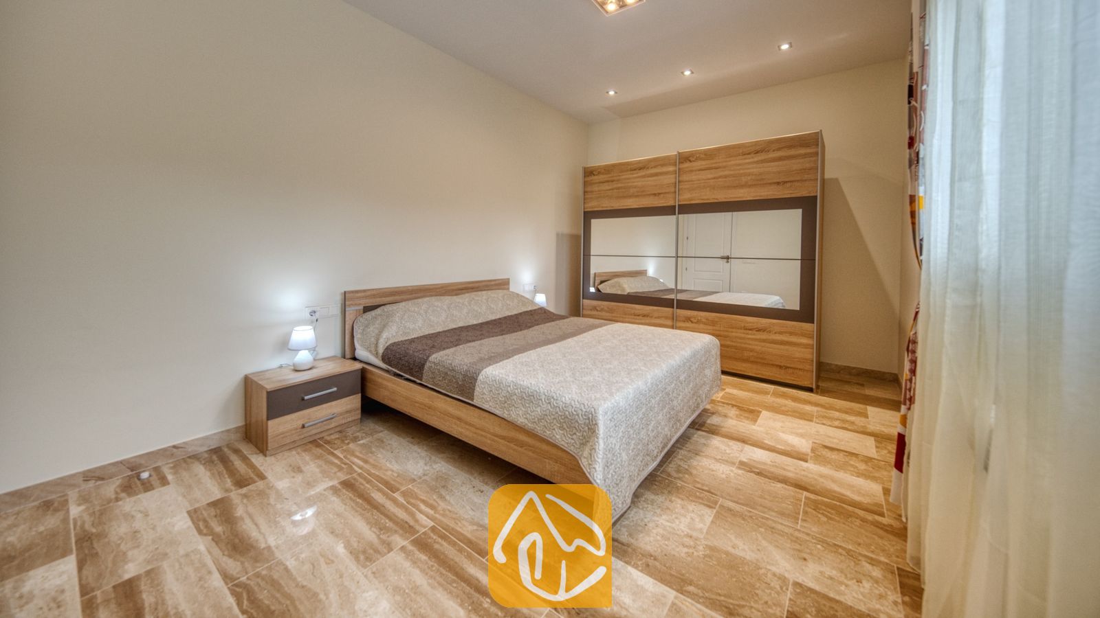Vakantievilla's Costa Brava Spanje - Villa Madison - Slaapkamer met een houten bedframe, een gedessineerd beige dekbed en een bijpassend nachtkastje met een klein lampje op een tegelvloer.