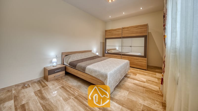 Holiday villas Costa Brava Spain - Villa Madison - Bedroom with a wooden bed frame, patterned beige duvet, and matching nightstand with a small lamp on tiled floors.