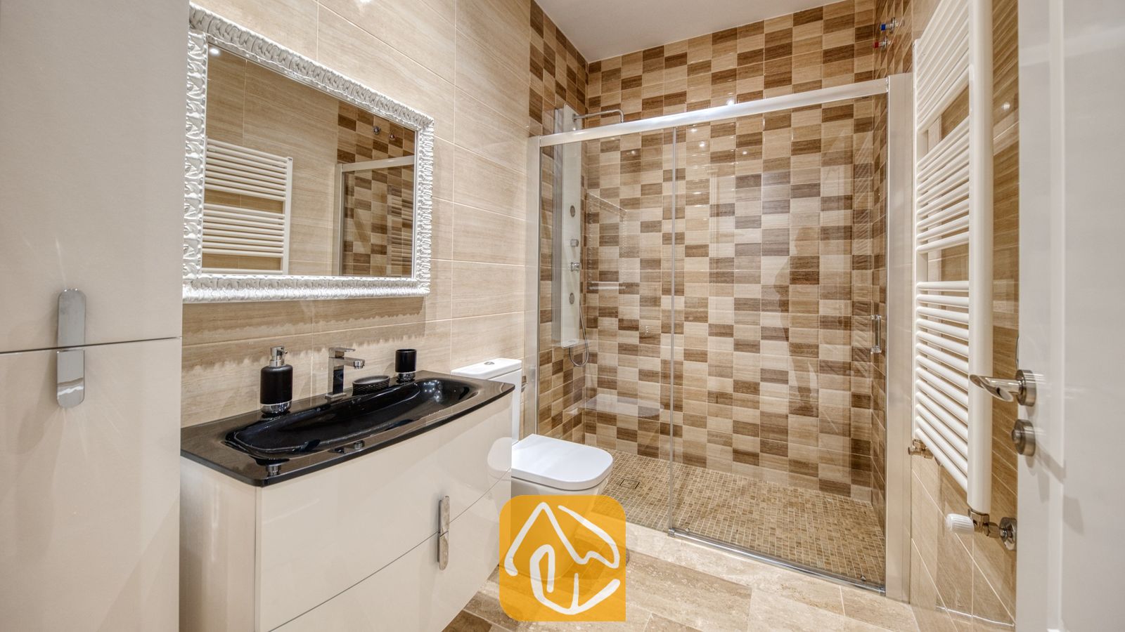 Vakantievilla's Costa Brava Spanje - Villa Madison - Moderne badkamer met een zwart wastafelmeubel, een glazen inloopdouche met bruine ruitjes tegels en een sierlijke spiegel met een wit lijstwerk.