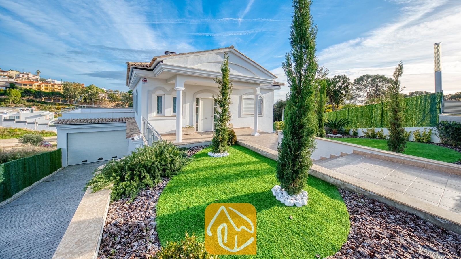 Vakantievilla's Costa Brava Spanje - Villa Madison - Buitenkant van Villa Madison met de witte gevel, hoge cipressen, een groene grasmat en een aangebouwde garage onder een blauwe lucht.