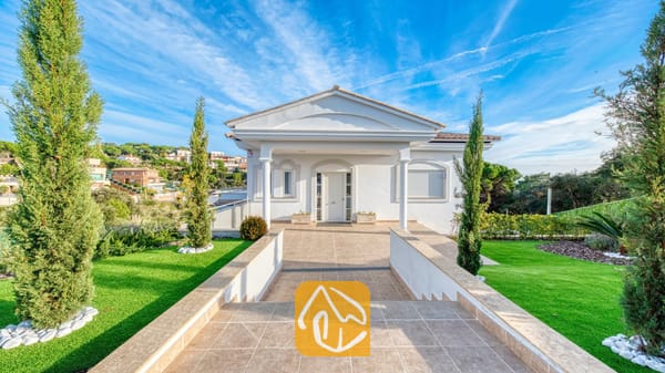 Vakantievilla's Costa Brava Spanje - Villa Madison - De witte buitenkant van Villa Madison met een betegeld pad als entree, zuilen en flankerende groene gazons met cipressen onder een blauwe lucht.