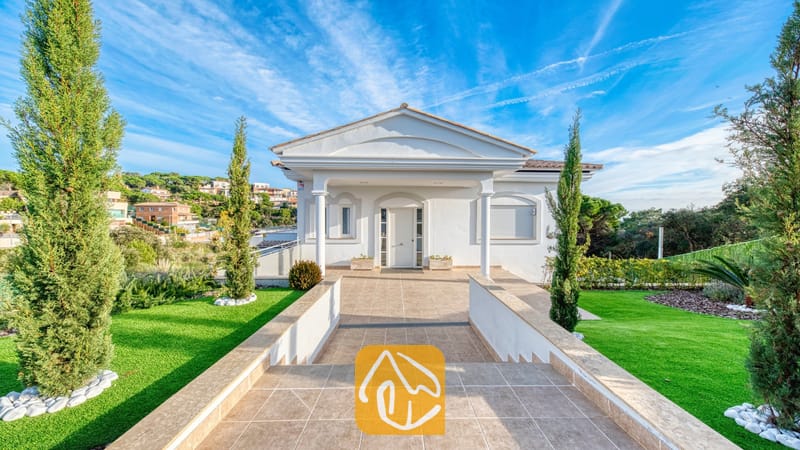 Vakantievilla's Costa Brava Spanje - Villa Madison - De witte buitenkant van Villa Madison met een betegeld pad als entree, zuilen en flankerende groene gazons met cipressen onder een blauwe lucht.