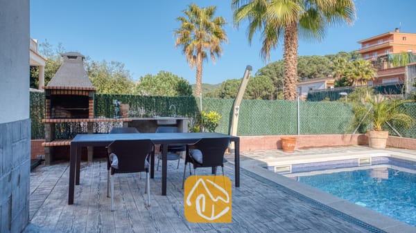 Vakantievilla's Costa Brava Spanje - Villa Zarita - Eetgedeelte buiten bij het zwembad van Villa Zarita, voorzien van een donkere Wicker tuintafel, vier stoelen, een ingebouwde stenen barbecue en palmbomen onder een blauwe lucht.